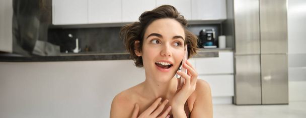 Close up portrait of young woman talking on mobile phone, looking surprised, hears amazing news, sitting near kitchen at home. Lifestyle and people concept - Photo, Image