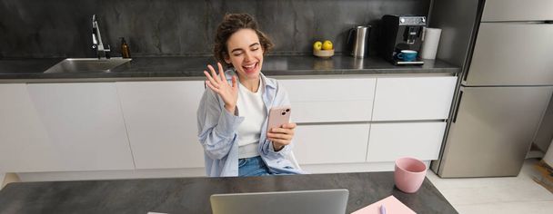 Πολυλογού μελαχρινή γυναίκα κάθεται στο σπίτι, βίντεο συνομιλίες με φίλο, κύματα χέρι στην κάμερα, κατέχει smartphone και χρησιμοποιεί φορητό υπολογιστή. - Φωτογραφία, εικόνα