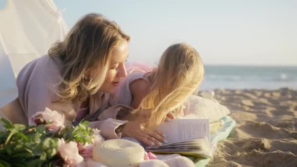 Happy parenthood concept. Preschooler kid daughter reading book having fun at carefree childhood. Beautiful loving family with blonde mother and little girl bonding at ocean beach during golden sunset - Footage, Video
