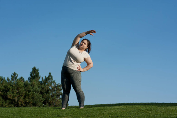 надмірна вага жінка середнього віку робить вправи в парку, стоячи на тлі трави, блакитного неба. жирна жінка нахиляє тіло на бік. Здоровий спосіб життя, позитивність тіла, пілатес на відкритому повітрі влітку - Фото, зображення