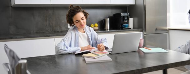 Egy kép fiatal nőről, aki kezeli a számláit, otthonról dolgozik laptoppal, leírja az információkat papírra. A tanuló házi feladatot csinál a konyhájában.. - Fotó, kép