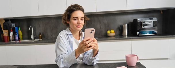 スマートフォンで自宅に座っている美しい笑顔の女性の肖像画,携帯電話アプリを使用して,注文食料品配達. - 写真・画像