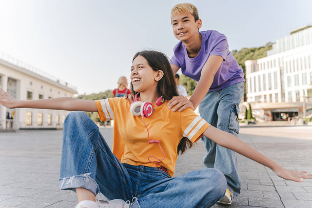 Happy teenage friends sitting, riding on skateboard on urban street. Group of happy multiracial boys and girls having fun together. School vacation, summer, friendship, positive lifestyle concept - Photo, Image