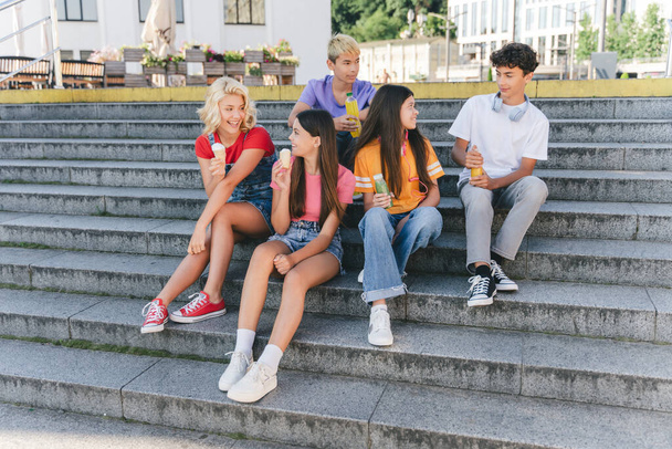 Grupa uśmiechniętych przyjaciół, wielorasowe nastolatki jedzące lody, pijące lemoniadę siedzącą na schodach. Szczęśliwy stylowy chłopców i dziewcząt komunikacji na ulicy. Przyjaźń, pozytywny styl życia, lato  - Zdjęcie, obraz
