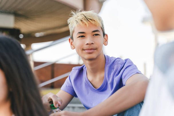 Portrét stylového usměvavého dospívajícího chlapce s barevnými vlasy, držícího láhev s pitím a dívajícího se stranou, sedícího s přáteli na městské ulici. Mládež, letní koncept  - Fotografie, Obrázek