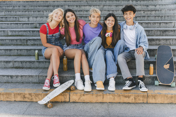 Groupe d'amis souriants, adolescents multiraciaux élégants assis sur des escaliers avec des planches à roulettes. Joyeux garçons et filles élégants communication, détente dans la rue. Amitié, mode de vie positif, été  - Photo, image