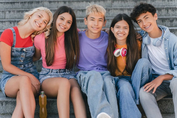 Grupo de amigos sorridentes, adolescentes multirraciais elegantes sentados nas escadas olhando para a câmera. Comunicação de meninos e meninas felizes, relaxando na rua. Amizade, estilo de vida positivo, verão  - Foto, Imagem