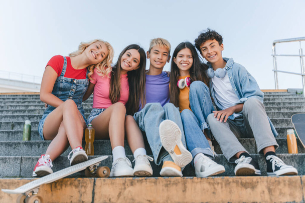 Retrato de adolescentes multirraciais sorridentes sentados em escadas olhando para a câmera na rua urbana. Amigos felizes abraçando juntos ao ar livre Amizade, conceito de diversidade  - Foto, Imagem