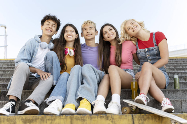 Grupa uśmiechniętych przyjaciół, wielorasowych nastolatków w kolorowych koszulkach przytulających się, siedzących na schodach na świeżym powietrzu. Szczęśliwych stylowych chłopców i dziewcząt na ulicy. Przyjaźń, pozytywny styl życia, pojęcie lata - Zdjęcie, obraz