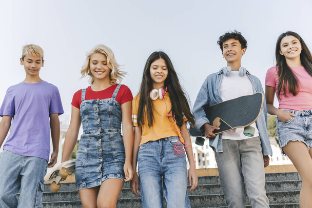 Grupo de amigos adolescentes multirraciais sorridentes vestindo camisetas coloridas conversando, comunicação, segurando skates andando na rua. Meninos e meninas elegantes felizes ao ar livre. Conceito de amizade  - Foto, Imagem