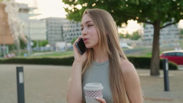 Relajada dama bebe café caliente teniendo conversación con su novio por teléfono. Mujer joven se ríe llamando novio en el parque de la ciudad de verano - Metraje, vídeo