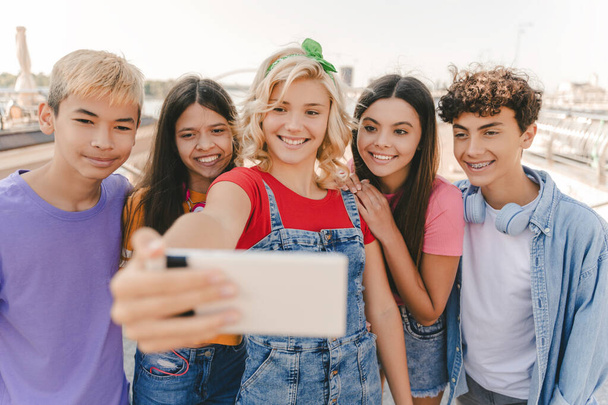 Grupa uśmiechniętych, wielorasowych nastolatków robiących selfie na ulicy. Młody blogerka influencer nagrywa wideo stojąc razem z przyjaciółmi. Technologia. Lato, pozytywny styl życia koncepcja  - Zdjęcie, obraz