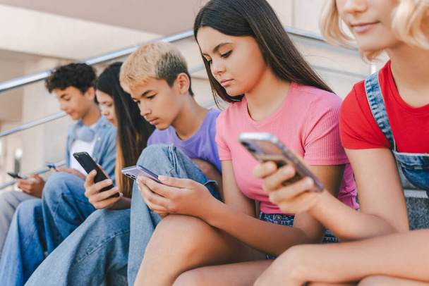 Bir grup ciddi çok ırklı genç ellerinde cep telefonları, video izliyor, internette iletişim kuruyor ve sokakta oturuyorlar. Teknoloji bağımlılığı kavramı  - Fotoğraf, Görsel