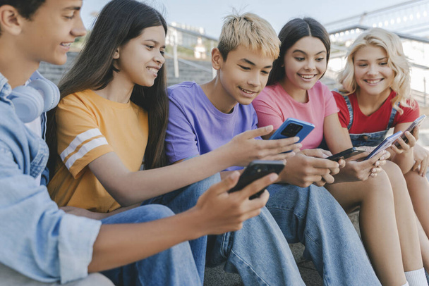 Group of smiling friends, multiracial teenagers holding mobile phones watching video, communication online, playing mobile game, talking sitting on stairs. Technology, social media concept - Photo, Image