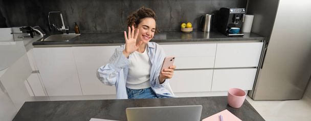 Молодая дружелюбная женщина, волны руку на ноутбук камеры, видео-чаты, держит смартфон, имеет беседу в Интернете. - Фото, изображение