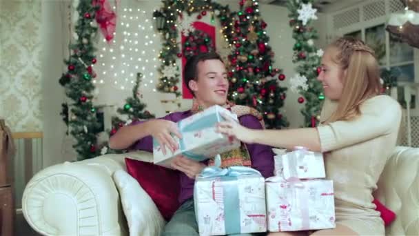 Szerető pár öltözött pulóvert és sálat gazdaság ajándékokat egy karácsonyi berendezett szobában. Fiatal pár szerelmes ember, és csinos lány visel pulóvert és sálat otthon. - Felvétel, videó