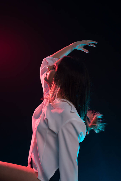 Young beautiful Caucasian woman sitting wearing white clothes and throwing her hair back with her right hand. Isolated on black background. - Photo, Image