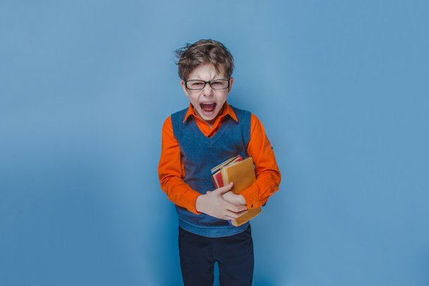 Menino de aparência europeia de dez anos de óculos abriu a boca gritando segurando um livro em um fundo azul
 - Foto, Imagem