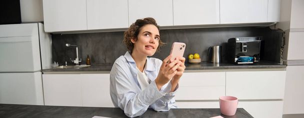 Portrait of woman thinking while holding smartphone, deciding what to order on mobile phone app. - Foto, Bild