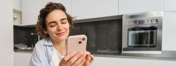 Portrait of modern brunette woman holding mobile phone and smiling. Girl sits in kitchen at home with smartphone, using app to order delivery, does online shopping. - Photo, Image