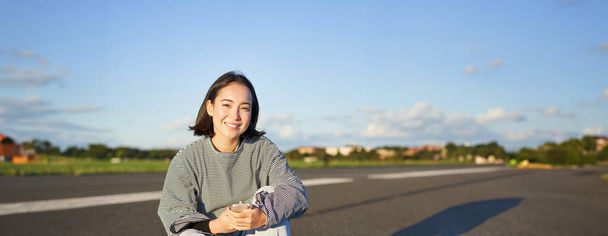 Plan vertical d'une femme asiatique assise sur une planche à roulettes sur la route, tenant une application smartphone. Skater fille skate sur longboard, en utilisant un téléphone mobile. - Photo, image