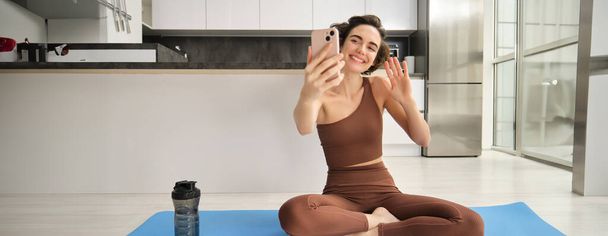 Retrato de una joven mujer de fitness, sentada en casa en una alfombra de goma, graba video en un teléfono inteligente, haciendo ejercicio vlog, haciendo sesión de entrenamiento en línea en vivo desde casa. Copiar espacio - Foto, Imagen