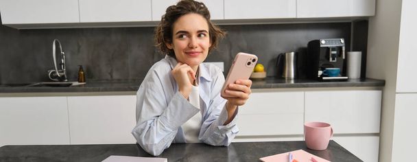 Portrait of beautiful smiling woman sitting at home with smartphone, using mobile phone app, orders groceries delivery. - Photo, Image