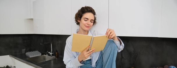Portrait of young modern woman sitting in kitchen, flipping pages, reading journal book, smiling happily. People and lifestyle concept - Фото, изображение