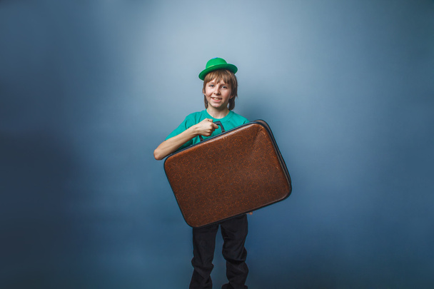European-looking boy of ten years in a hat holding a suitcase in hand, itinerary on a blue background - Fotó, kép