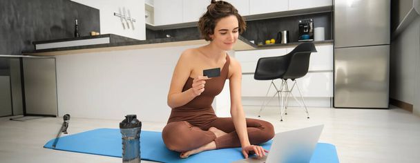 Retrato de atleta joven, chica de yoga pagando por clases en línea, comprando entrenamiento remoto con instructor de gimnasio, sentada en su estera de goma con computadora portátil y tarjeta de crédito. - Foto, imagen