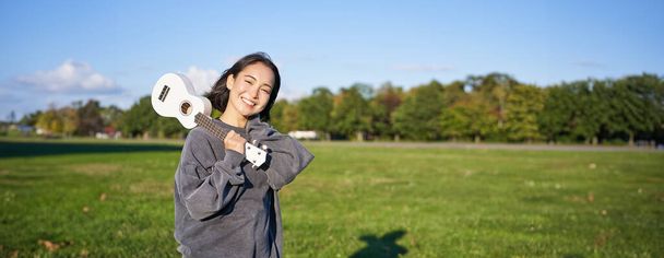 Portret van een prachtig lachend meisje met ukelele, aziatische vrouw met muziekinstrument dat buiten poseert in het groene park. Mensen en levensstijl - Foto, afbeelding