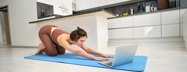 Imagen de la chica atleta, deportista practica yoga, siga la videoclase en línea en el portátil, viendo las instrucciones de entrenamiento en el ordenador, haciendo ejercicio desde casa. - Foto, imagen