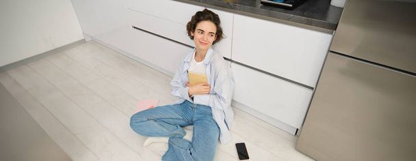 Άνω γωνία πλάνο της νεαρής γυναίκας που κάθεται στο πάτωμα της κουζίνας, αναθεώρηση για εξετάσεις, διαβάζοντας τις σημειώσεις της, κάνει την εργασία στο σπίτι. - Φωτογραφία, εικόνα