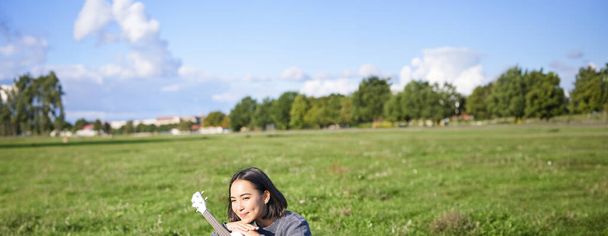 Ρομαντικό κορίτσι από την Ασία κάθεται με ukulele κιθάρα στο πάρκο και χαμογελώντας, χαλαρώνοντας μετά το πανεπιστήμιο, απολαμβάνοντας ρεπό στον καθαρό αέρα. - Φωτογραφία, εικόνα