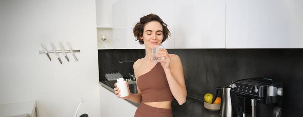 Portrait of fitness girl, healthy woman standing in kitchen with pills, takes vitamins with glass of water for healthy wellbeing. Sportswoman holds food supplements. - Фото, изображение