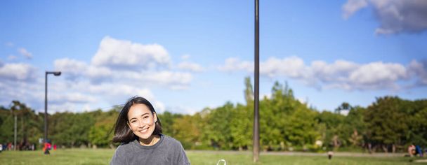Vertical shot of cute teen girl sits in park on grass with backpack and her book, reading alone outdoors. - Foto, afbeelding