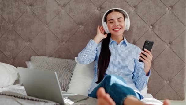 Joyful lady with dark long hair listening music in wireless headphones holding mobile phone in hand with favorite track. Female manager tourist having fun sitting on bed hotel relax after work. - Footage, Video