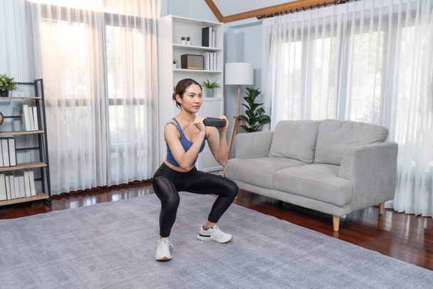 Vigorous energetic woman doing squat weight lifting exercise at home. Young athletic asian woman strength and endurance training session as home workout routine with squat. - Photo, Image