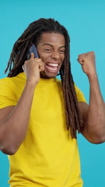 Latin man with dreadlocks celebrating while talking to the mobile in studio with blue background - Footage, Video