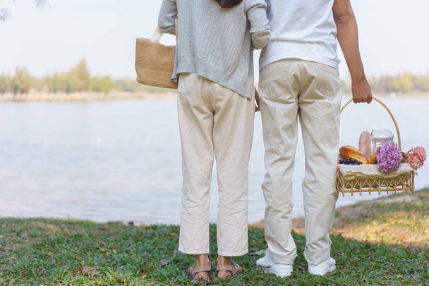Νεαρό ζευγάρι μεταφέρουν καλάθι πικ-νικ και αγκαλιάζει μαζί, ενώ στέκεται κοντά στη λίμνη στο πάρκο. - Φωτογραφία, εικόνα