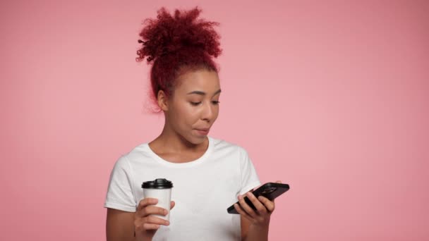 Közelkép mosolygós afro-amerikai üzletasszony gépelés böngészés okostelefon tartja csésze kávé vagy tea a kezében. Vöröshajú nő dolgozik mobiltelefon online távoli izolált rózsaszín háttér - Felvétel, videó