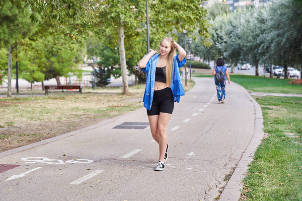 Fiatal, szőke hajú kaukázusi nő sétál a szabadban egy parkban, inline görkorcsolyával a kezében, hogy korcsolyázni menjen, és sportoljon egy forró, napos napon. - Fotó, kép