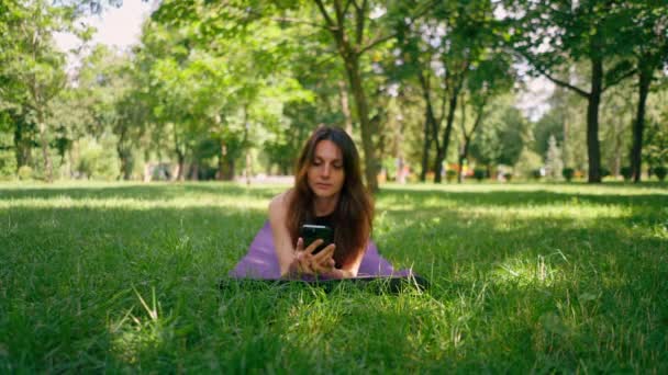 Aktiivinen kaunis joogi nainen kävelyllä puistossa ennen tai jälkeen koulutus seisoo kunto matto tilalla älypuhelin käsissään - Materiaali, video