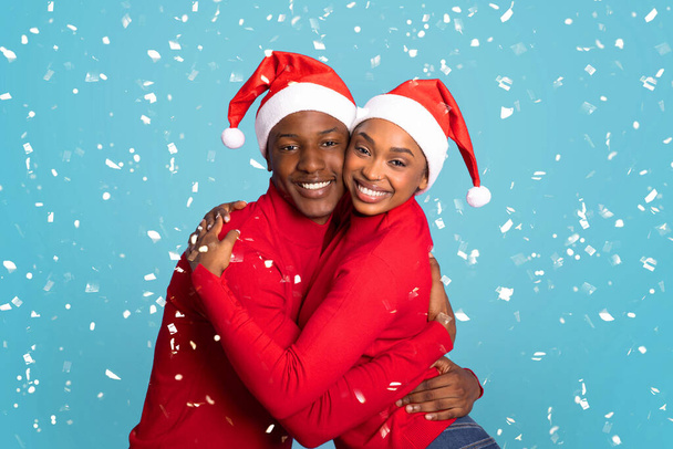 Weihnachtsstimmung. Glückliches schwarzes Paar mit Weihnachtsmützen, die sich unter fallenden Konfetti umarmen, lächelnd in die Kamera auf blauem Studiohintergrund. Weihnachtsfeier, Konzept für die Winterferien - Foto, Bild