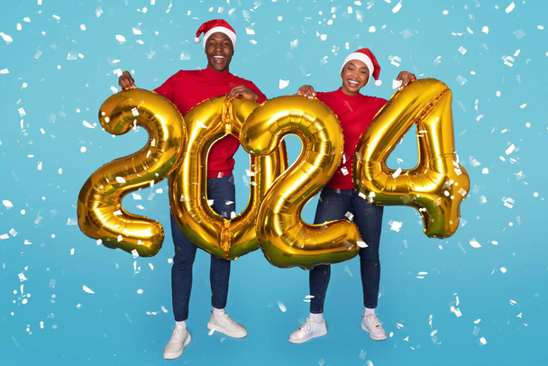 Vrolijk Afro-Amerikaans paar Holding Golden 2024 ballonnen staande op turkoois blauwe achtergrond, vieren komende nieuwjaar te midden van dalende confetti, het dragen van Santa hoeden. Volledige lengte schot - Foto, afbeelding
