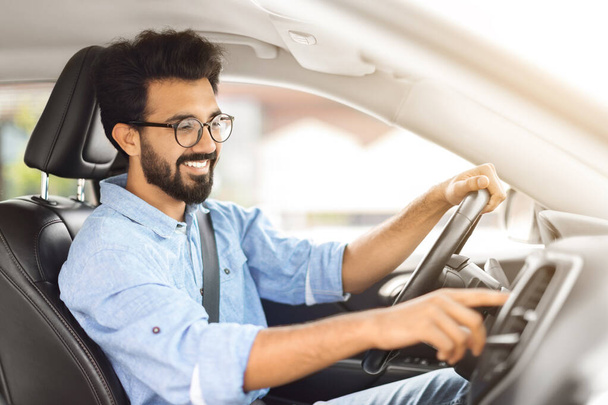 Drogowa playlista. Side Zobacz profil Portret Happy Indian Man Driving Car And Listening To Music On Auto Audio System, Sprawdzanie Opcje samochodowe podczas jazdy testowej Wciskanie przycisku Włączanie radia - Zdjęcie, obraz