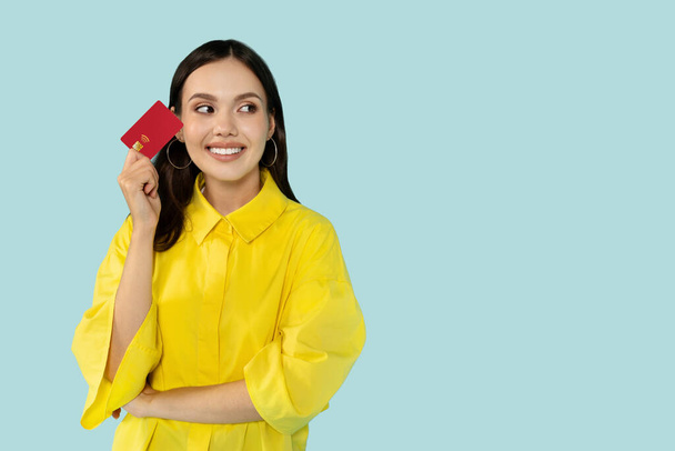 Čas nakupovat. Snící mladá bruneta žena ve žluté košili drží kreditní kartu a dívá se jinam, pózuje přes modré studio pozadí, panorama s kopírovacím prostorem - Fotografie, Obrázek