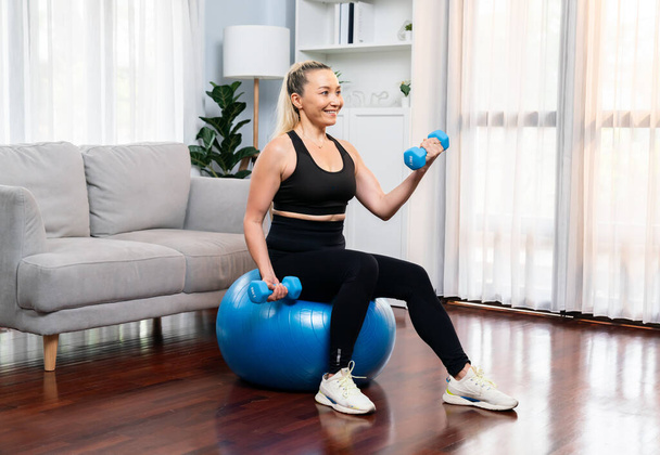 Athletic and sporty senior woman sitting on fit ball while engaging in weight lifting with dumbbell at home exercise as concept of healthy fit body lifestyle after retirement. Clout - Photo, Image