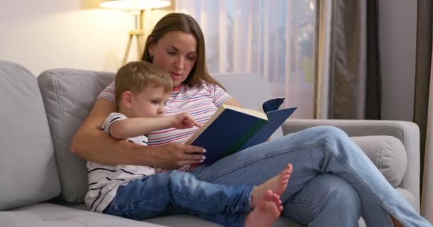 Moeder en zoon lezen een boek zittend op een gezellige bank in de woonkamer, familietijd. Hoge kwaliteit 4k beeldmateriaal - Video