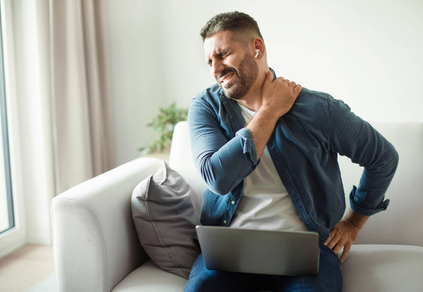 Middle Aged European Man Suffering From Neckache And Backache After Online Work On Laptop Computer, Sitting On Sofa At Home. Sick Guy With Earbuds Rubbing Body Inflamed Zones, Feeling Unwell - Photo, Image